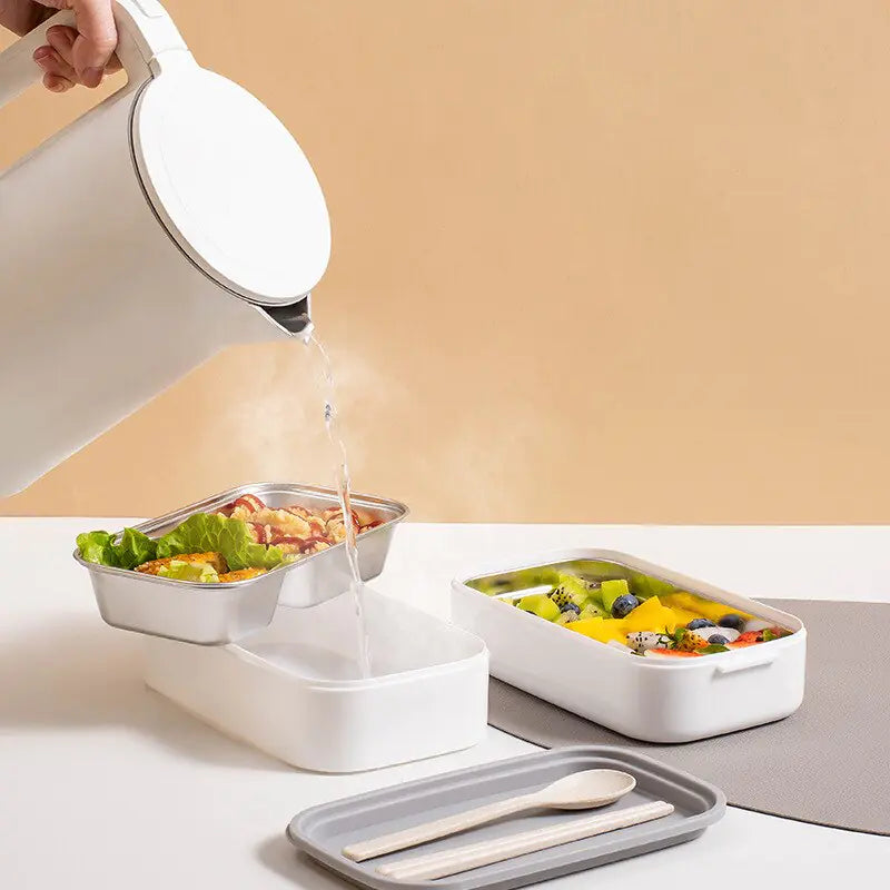 Weiße Bento-Box aus Kunststoff mit Fächern, gefüllt mit Salat und Obst, auf Tisch.