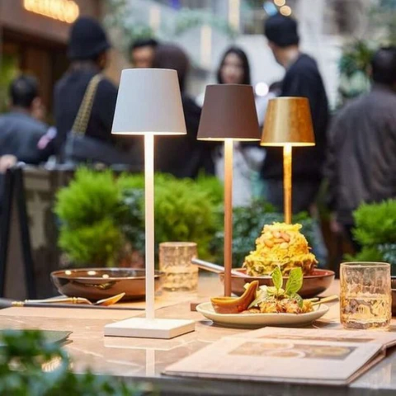 Drei moderne Tischlampen in Weiß, Schwarz und Gold auf einem Restauranttisch mit Essen und Getränken.