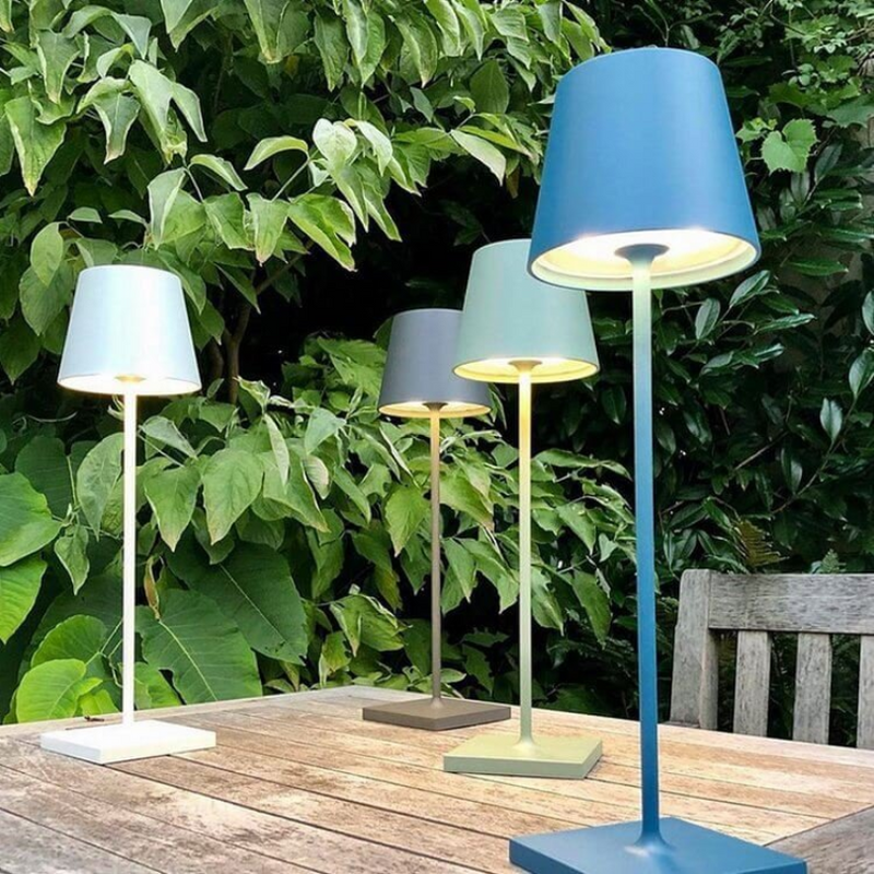 Vier bunte Tischlampen auf Holztisch im Garten, moderne Außenbeleuchtung, grüne Blätter im Hintergrund.