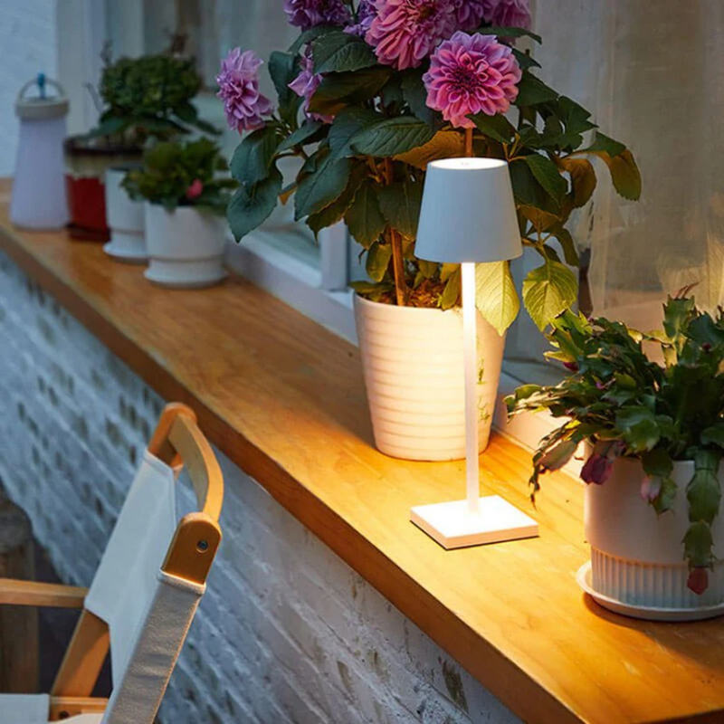 Weiße Tischlampe auf Holzfensterbank mit blühenden Pflanzen, ideal für stimmungsvolle Beleuchtung.