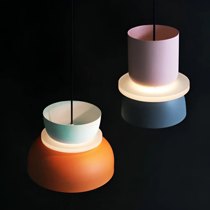 Zwei moderne Hängelampen, orange und rosa, aus Metall, beleuchtet, vor schwarzem Hintergrund.