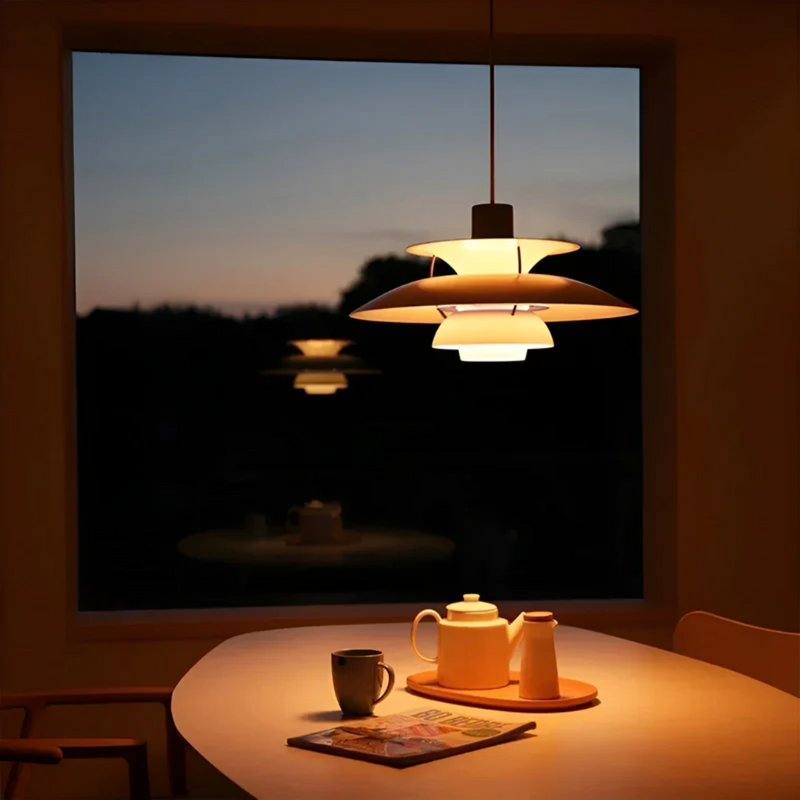 Moderne Hängelampe über Esstisch, beleuchtet, mit Teekanne und Tasse, Abenddämmerung im Hintergrund.