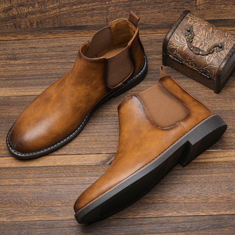 Braune Chelsea-Stiefel aus Leder auf Holzboden neben dekorativer Box.