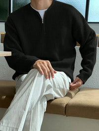 Mann in schwarzem Strickpullover und weißen Hosen sitzt auf Sofa, moderne Freizeitkleidung.