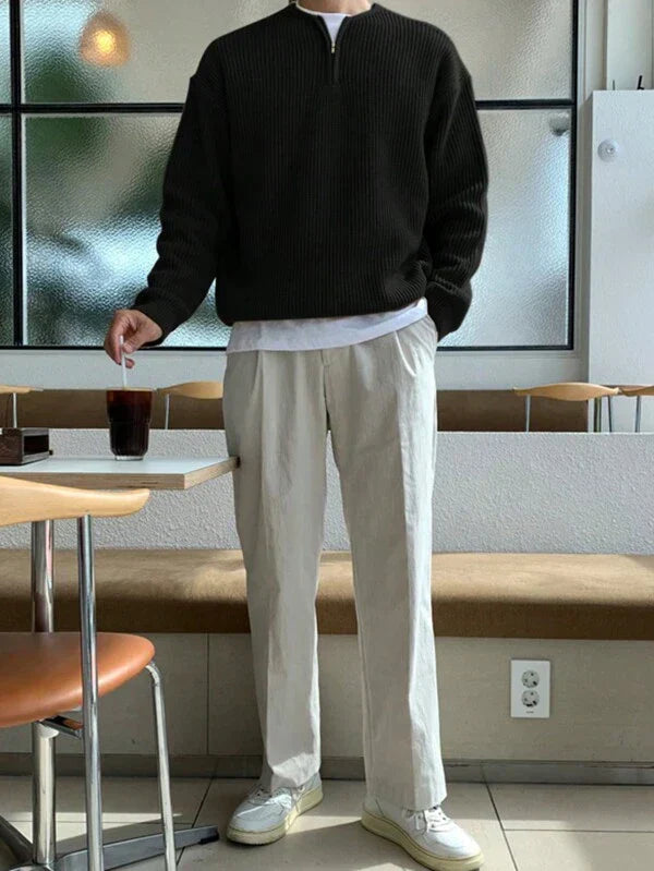 Mann in schwarzem Pullover und beigen Hosen, hält ein Getränk, sitzt in modernem Café.