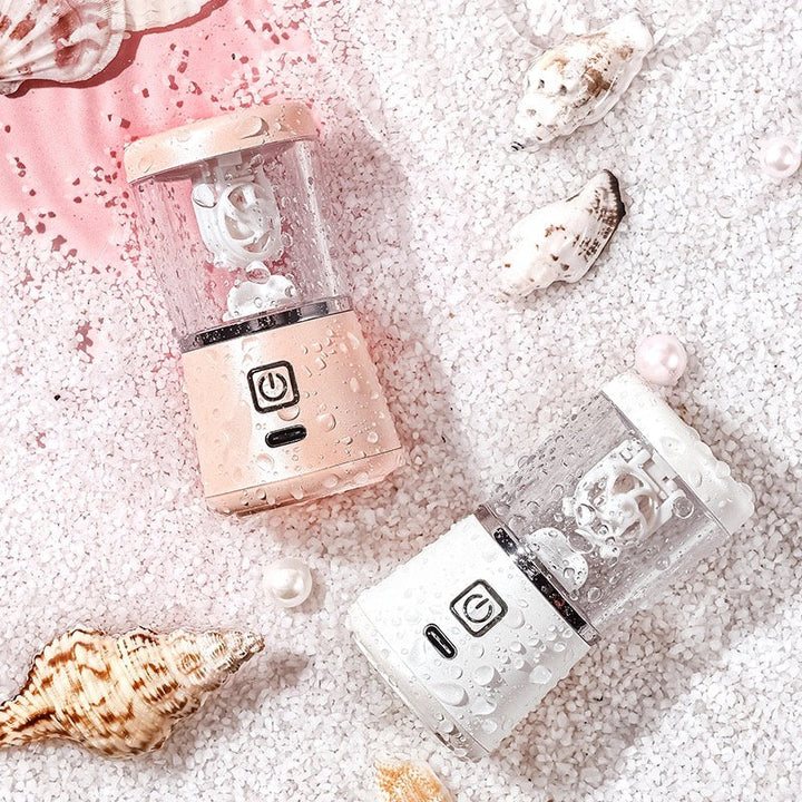 Zwei tragbare Mini-Mixer in Rosa und Weiß auf Sand mit Muscheln und Perlen.