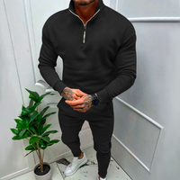 Mann in schwarzem Trainingsanzug mit Reißverschluss, weiße Sneaker, Zimmerpflanze im Hintergrund.
