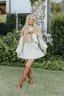Frau in cremefarbenem Strickkleid mit braunen Stiefeln im Garten, modisch, elegant, Herbstmode.
