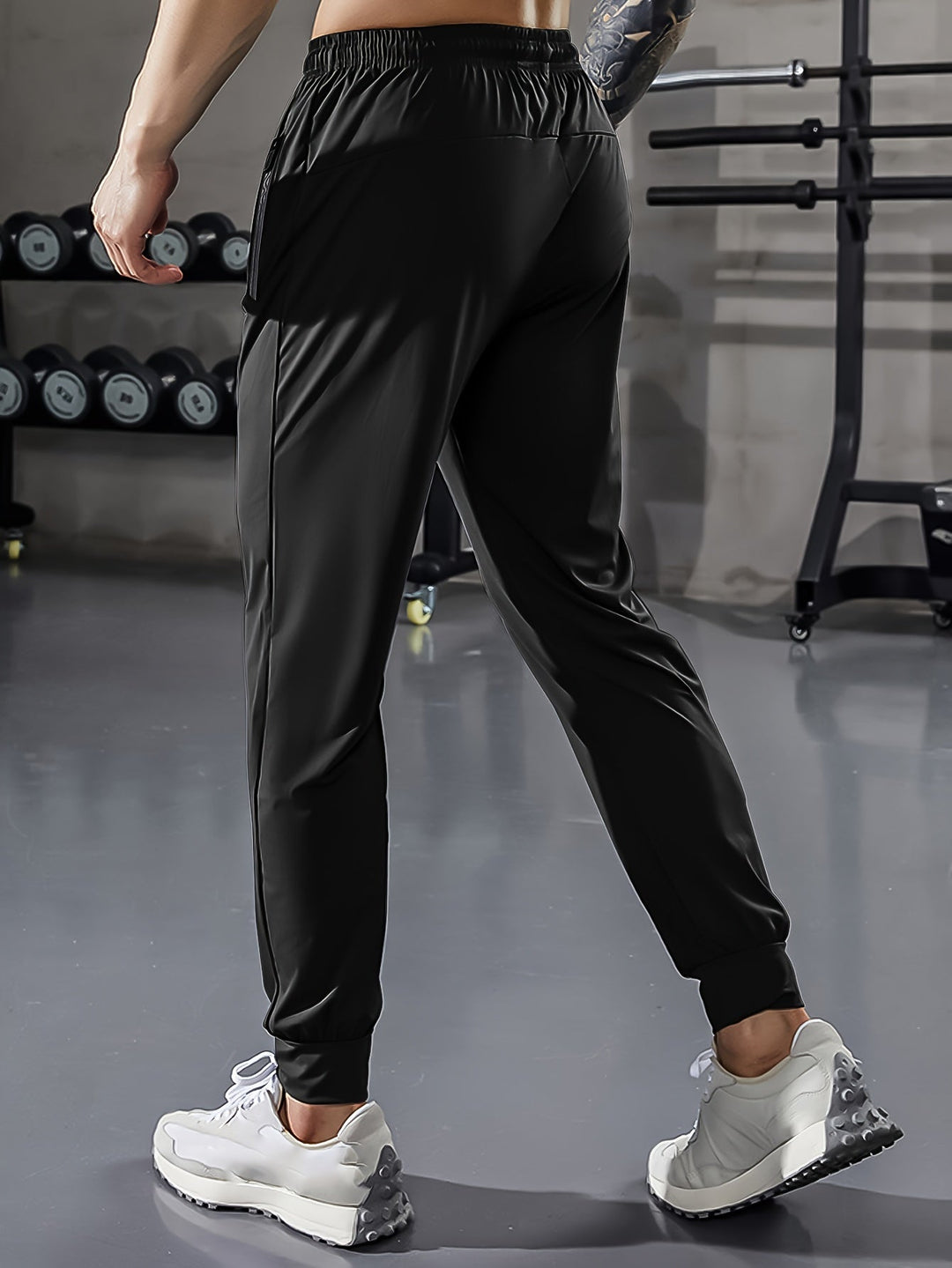 Mann in schwarzer Sporthose im Fitnessstudio, Hanteln im Hintergrund, weiße Turnschuhe.