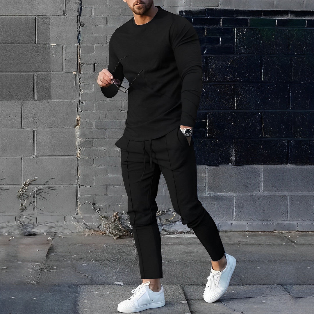 Mann in schwarzem Outfit mit weißen Sneakers vor grauer Ziegelwand, modisch, urbaner Stil.