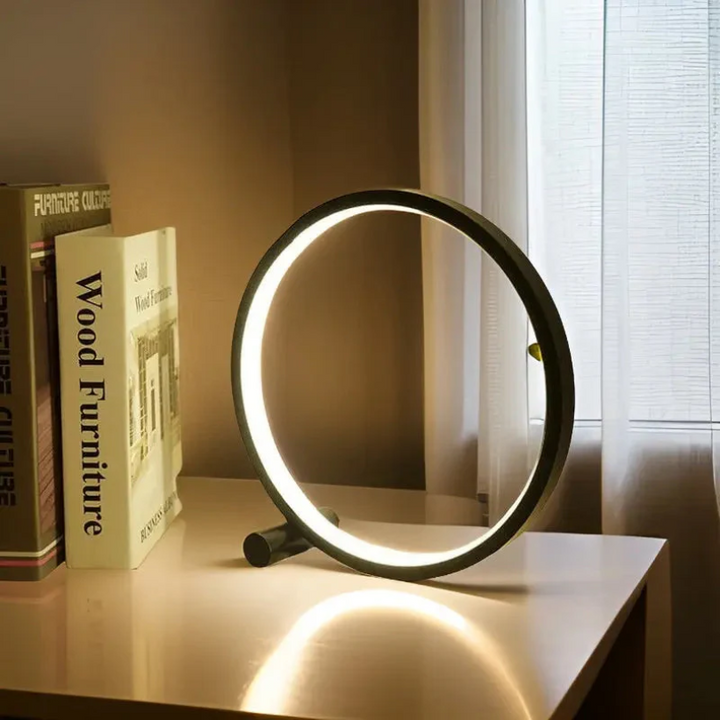 Moderne LED-Tischlampe, rund, schwarz, auf weißem Tisch neben Büchern und Fenster.