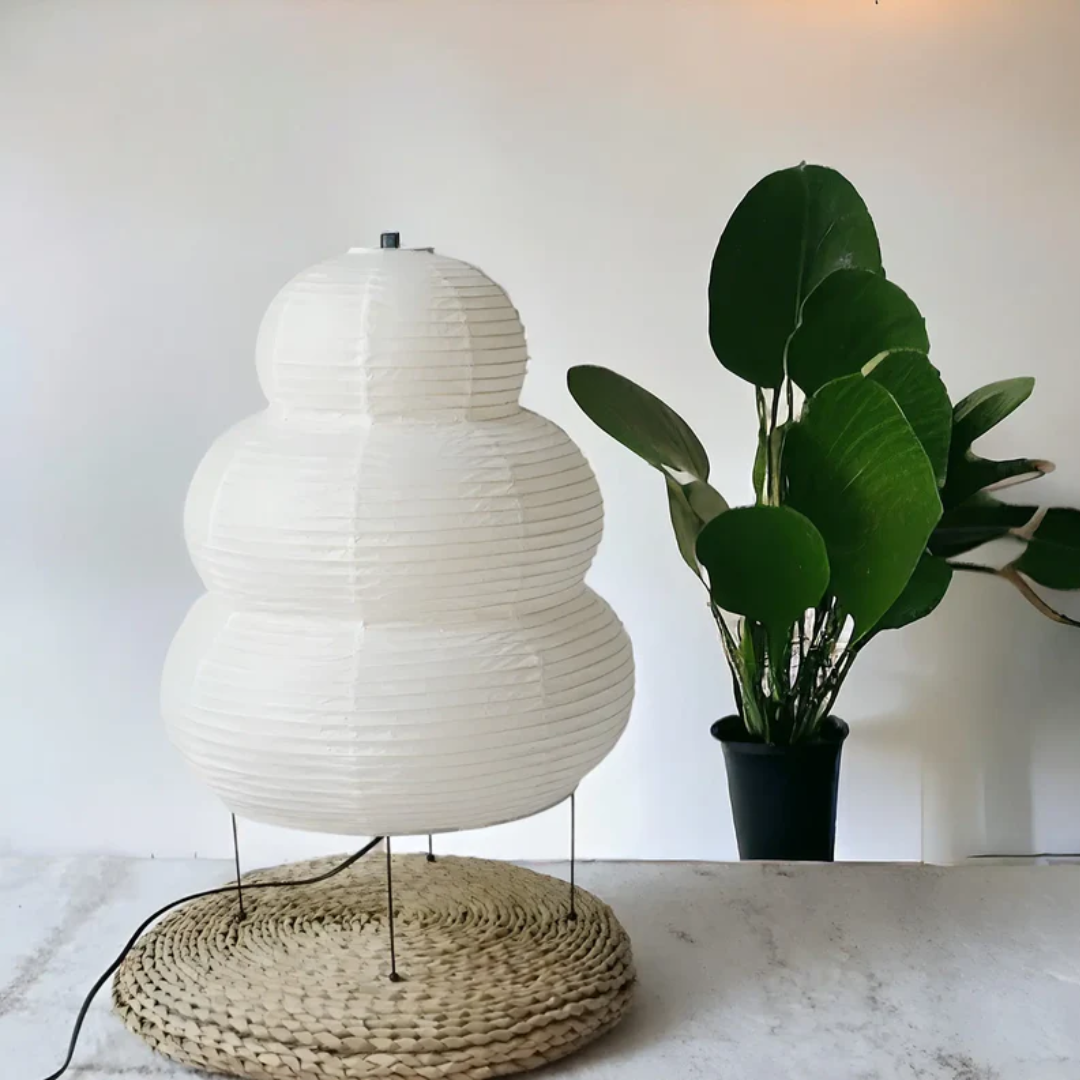 Weiße Papierlampe auf geflochtener Unterlage neben grünem Topfpflanze, modernes Innendesign.