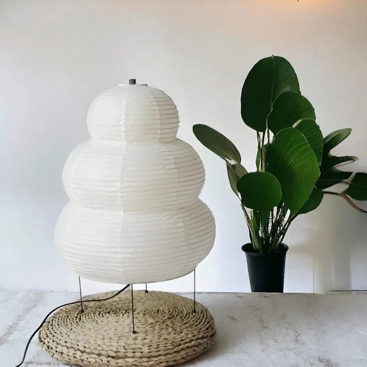 Weiße Papierlampe auf geflochtener Unterlage neben grünem Topfpflanze, modernes Innendesign.