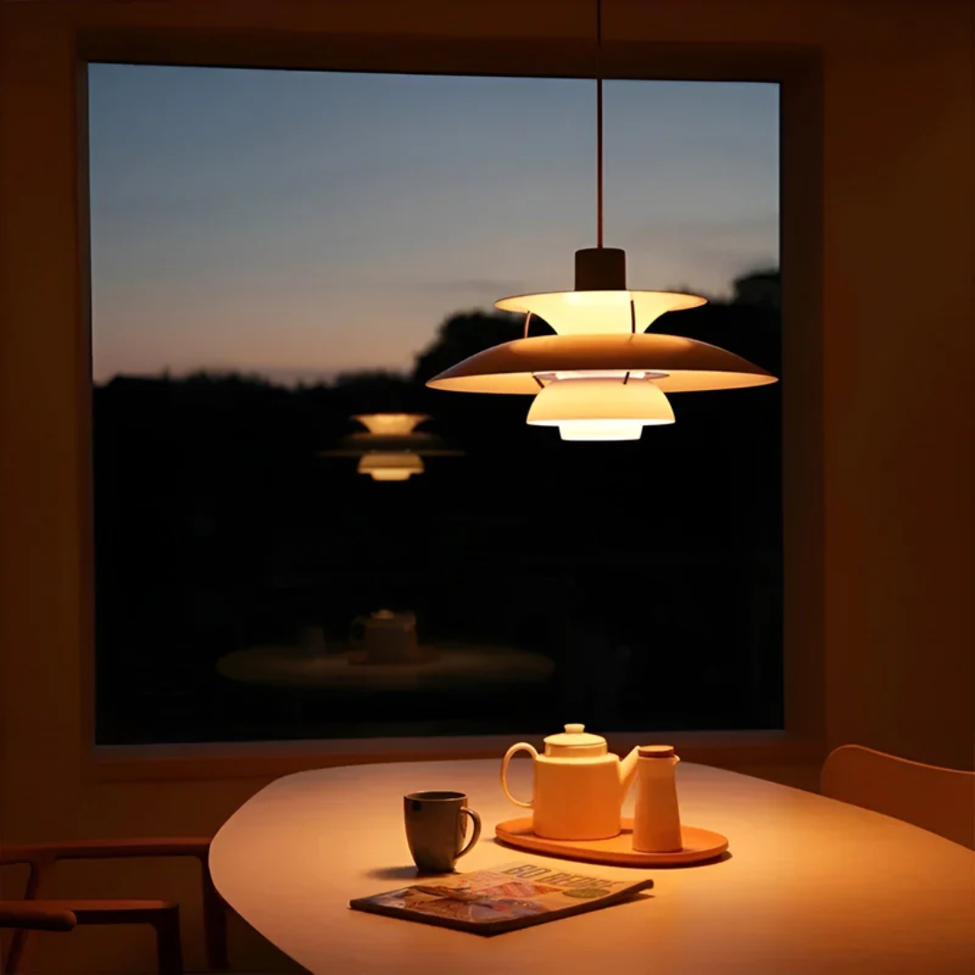 Moderne Hängelampe über Esstisch, beleuchtet, mit Teekanne und Tasse, Abenddämmerung im Hintergrund.