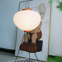 Moderne Stehlampe mit weißem Lampenschirm und schwarzem Stativ, vor Kunstwerk im Hintergrund.