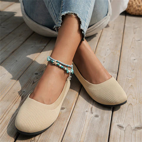 Beige Damen-Ballerinas aus Stoff, lässig, mit türkisfarbenem Fußkettchen, auf Holzboden.