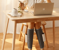 Frau nutzt grauen Beinmassagegerät unter Holztisch, modernes Design, Entspannung, Wellness.