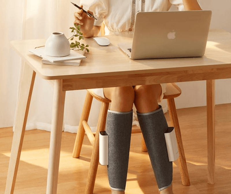 Frau nutzt grauen Beinmassagegerät unter Holztisch, modernes Design, Entspannung, Wellness.