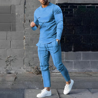 Mann in blauem Trainingsanzug, hält Sonnenbrille, weiße Sneaker, graue Ziegelwand im Hintergrund.