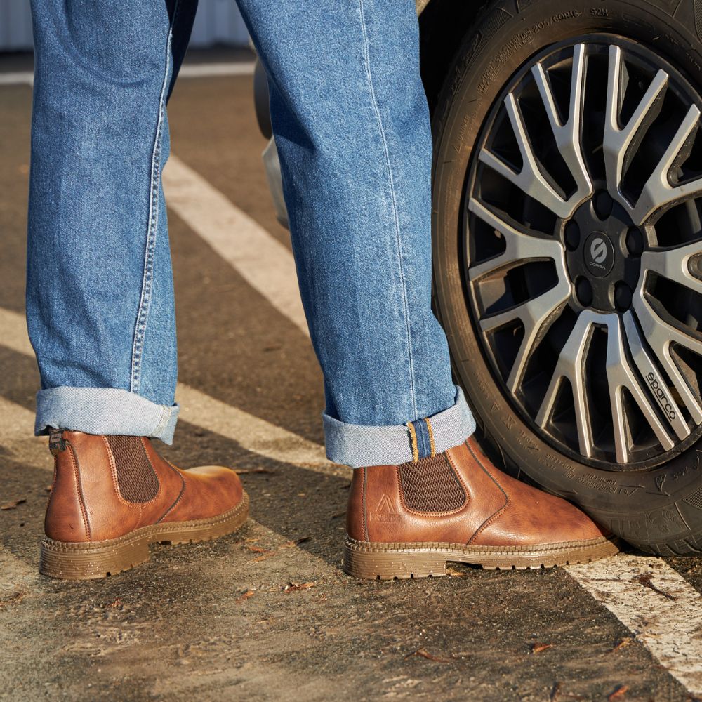 Braune Chelsea-Boots aus Leder, Herrenmode, neben Autoreifen, lässiger Stil, Jeans.