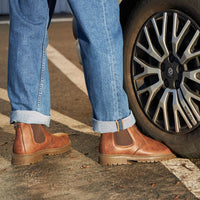 Braune Chelsea-Boots aus Leder, Herrenmode, neben Autoreifen, lässiger Stil, Jeans.