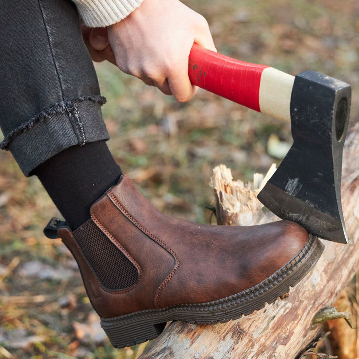 Person mit braunen Chelsea-Boots spaltet Holz mit rotem Axtstiel im Freien.