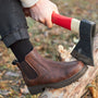 Person mit braunen Chelsea-Boots spaltet Holz mit rotem Axtstiel im Freien.