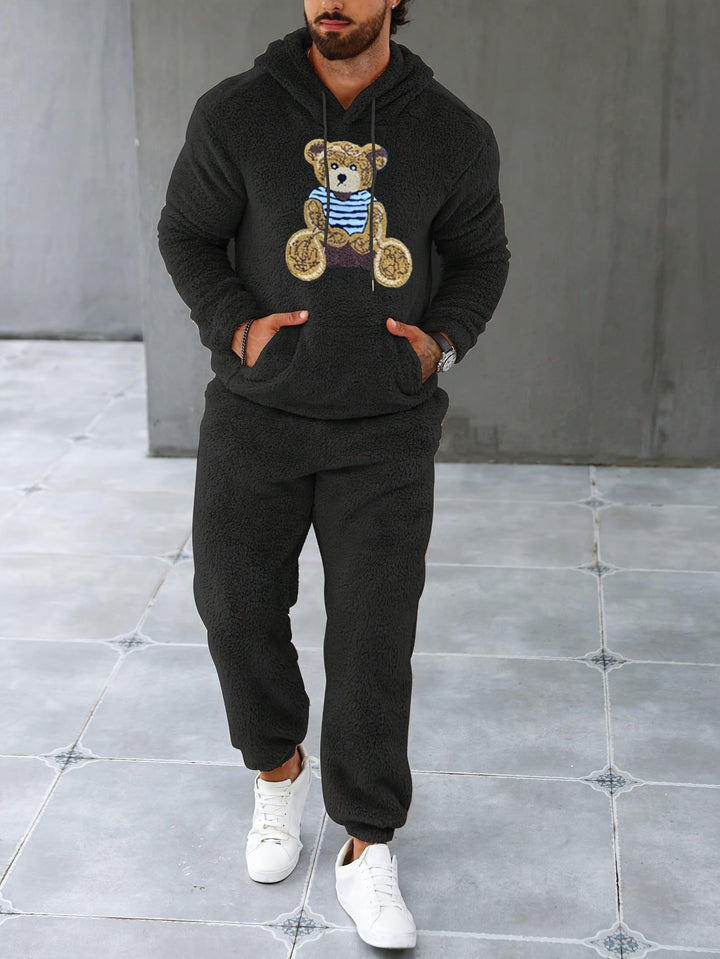 Mann in schwarzem Teddy-Fleece-Trainingsanzug mit Bärenmotiv, weiße Sneaker, grauer Hintergrund.