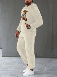 Mann in cremefarbenem Teddyfleece-Trainingsanzug mit Bärenmotiv, weiße Sneaker.