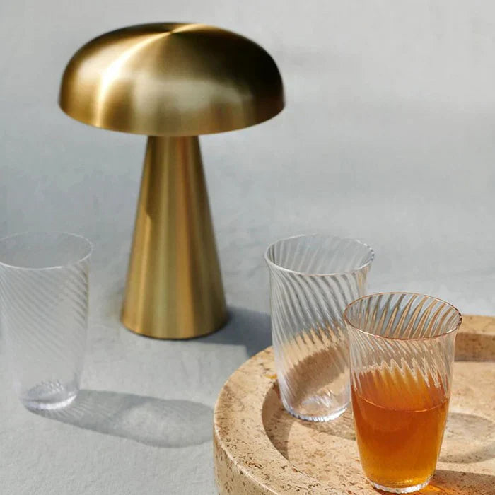 Goldene Tischlampe aus Metall neben gerippten Gläsern auf hellem Hintergrund.