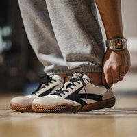 Person trägt beige Sneaker mit schwarzem Logo und brauner Sohle, graue Jogginghose, Armbanduhr.