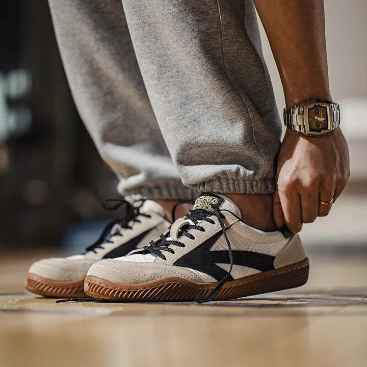 Person trägt beige Sneaker mit schwarzem Logo und brauner Sohle, graue Jogginghose, Armbanduhr.