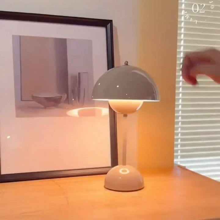 Graue Tischlampe aus Metall auf Holzoberfläche neben gerahmtem Bild und Fensterjalousie.