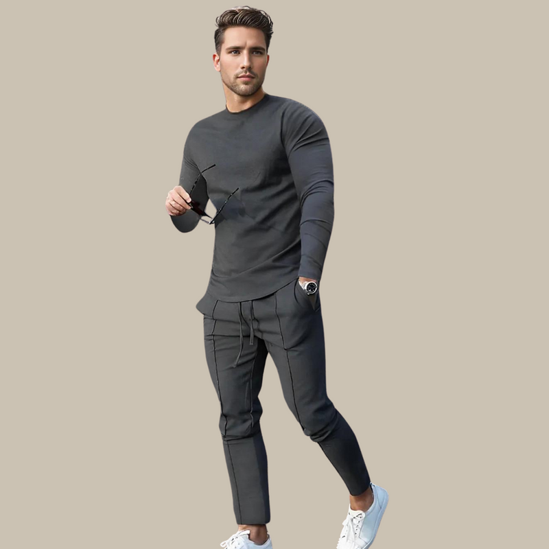 Mann in grauem Freizeitanzug mit Sonnenbrille und weißen Sneakers vor beigem Hintergrund.