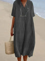 Frau in grauem Leinenkleid am Strand mit Strohtasche und Sonnenbrille.