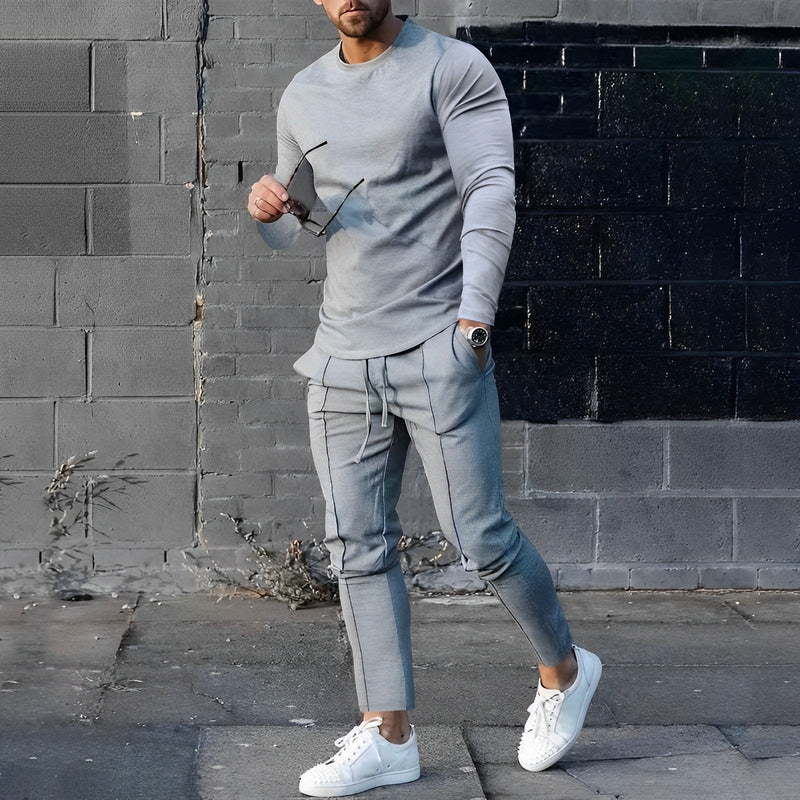 Mann in grauem Jogginganzug und weißen Sneakers vor schwarzer Ziegelwand.