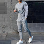 Mann in grauem Jogginganzug und weißen Sneakers vor schwarzer Ziegelwand.