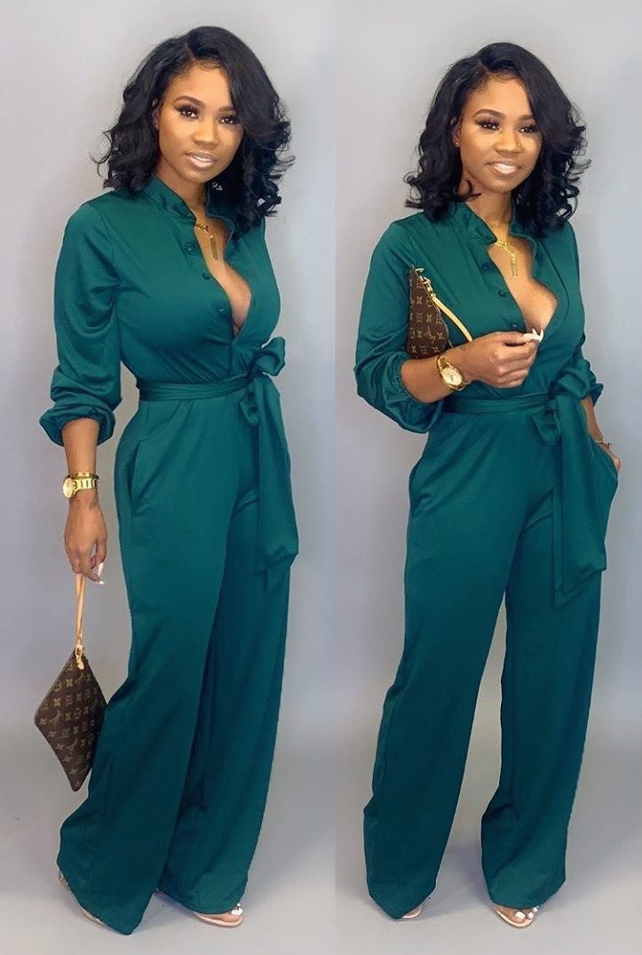 Frau in grünem Jumpsuit mit Gürtel, goldener Uhr und brauner Clutch vor grauem Hintergrund.