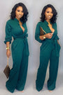 Frau in grünem Jumpsuit mit Gürtel, goldener Uhr und brauner Clutch vor grauem Hintergrund.