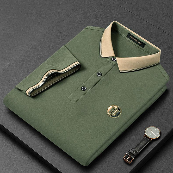 Grünes Herren-Poloshirt mit beigem Kragen, gefaltet, neben einer Armbanduhr auf grauem Hintergrund.