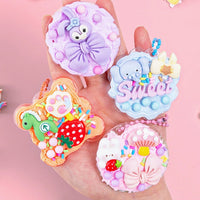 Hand hält bunte, dekorative Handy-Anhänger mit Tiermotiven und Süßigkeiten-Designs auf rosa Hintergrund.