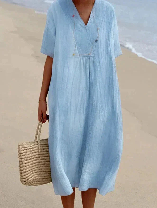 Frau in hellblauem Leinenkleid am Strand mit Strohtasche.