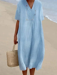 Frau in hellblauem Leinenkleid am Strand mit Strohtasche.