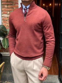 Mann in rotem Pullover mit Reißverschluss, beiger Hose, Krawatte, Armbanduhr, vor Backsteinwand.