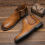 Braune Chelsea-Stiefel aus Leder auf Holzboden neben dekorativer Box.