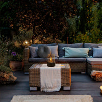 Rattan-Gartensofa mit blauen Kissen, Couchtisch, Tischfeuerstelle, abends im beleuchteten Garten.