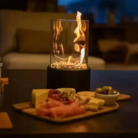 Tischkamin mit Glaszylinder, brennend, auf Holztisch neben Käseplatte und Oliven, gemütliche Atmosphäre.
