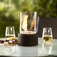 Glas-Feuerstelle auf Tisch, umgeben von Weingläsern, ideal für Garten oder Terrasse.