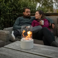 Tischkamin mit Glaszylinder, weißem Sockel, Flammen, Outdoor, gemütliche Atmosphäre, Terrasse.