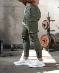 Mann in olivgrüner Jogginghose und weißen Sneakers im Fitnessstudio, Hantelbank im Hintergrund.